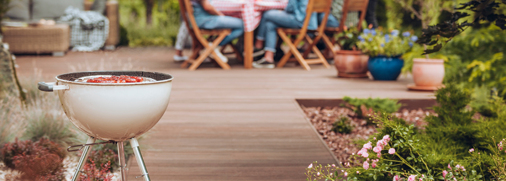 Grill mit Würstchen im Garten mit Blumen und Gruppe von Freunden, die im Hintergrund feiern